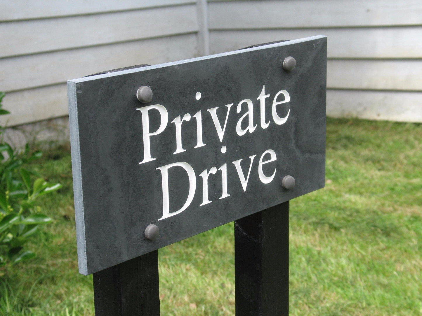 A slate house sign, measuring 200mm x 100mm, deep-engraved house name in Times New Roman font, on charcoal slate, enhanced with white paint. It is mounted on two wooden posts.