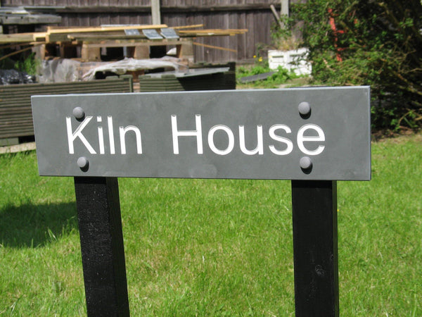 A slate house sign, measuring 300mm x 100mm, deep-engraved house name in Arial font, on grey slate, enhanced with white paint. It is mounted on two wooden post.
