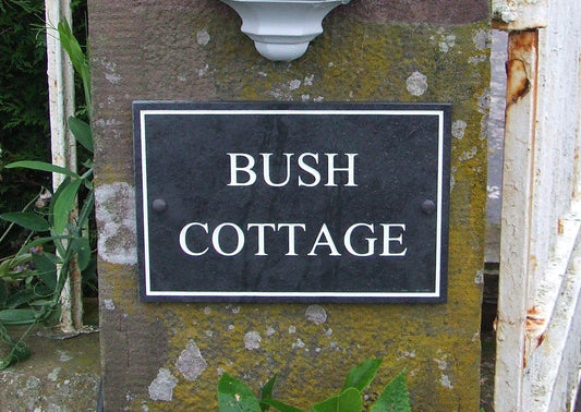 A slate house sign, measuring 400mm x 300mm with a plain border, engraved Bush Cottage in Times New Roman font, on grey slate, with white paint. 
